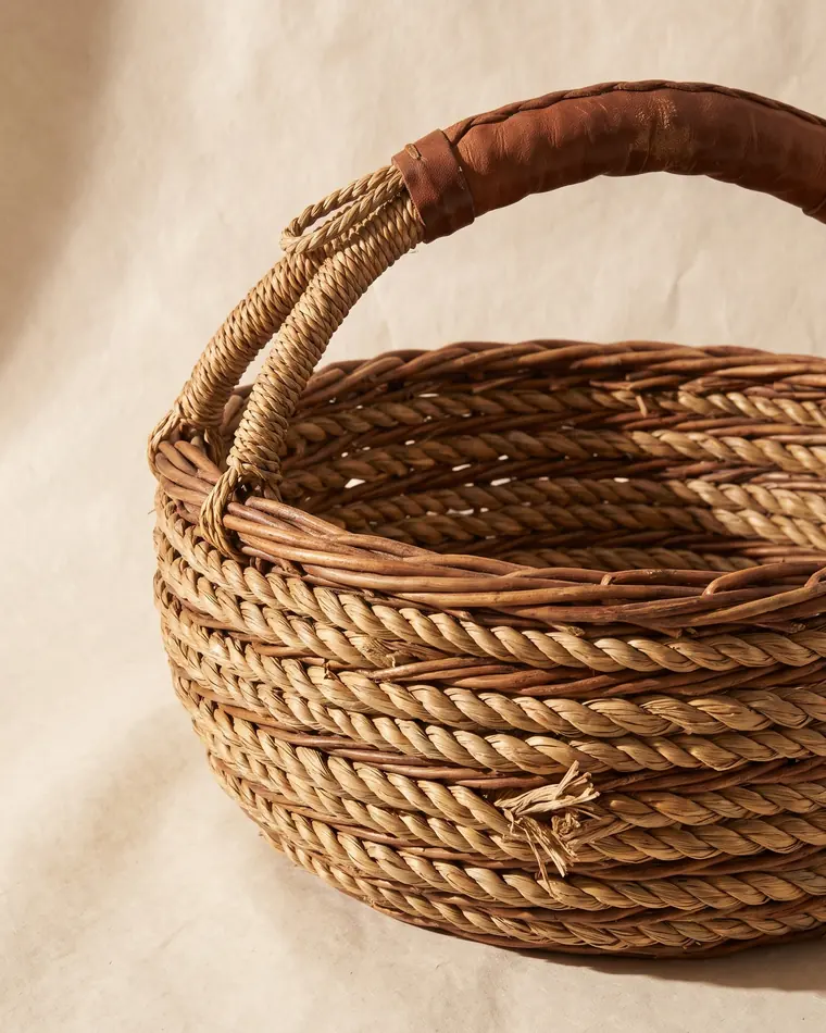 A large coiled willow and seagrass basket with a worn leather handle wrap on a warm paper sweep.