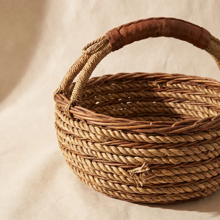 A large coiled willow gathering basket with a worn handle on a warm paper sweep — lower detail, showing the foot and the shadow.