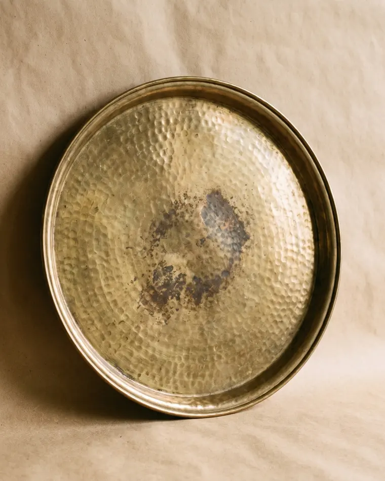 A large round hammered brass tray with a rolled rim and even tarnish, leaning on a warm paper sweep.