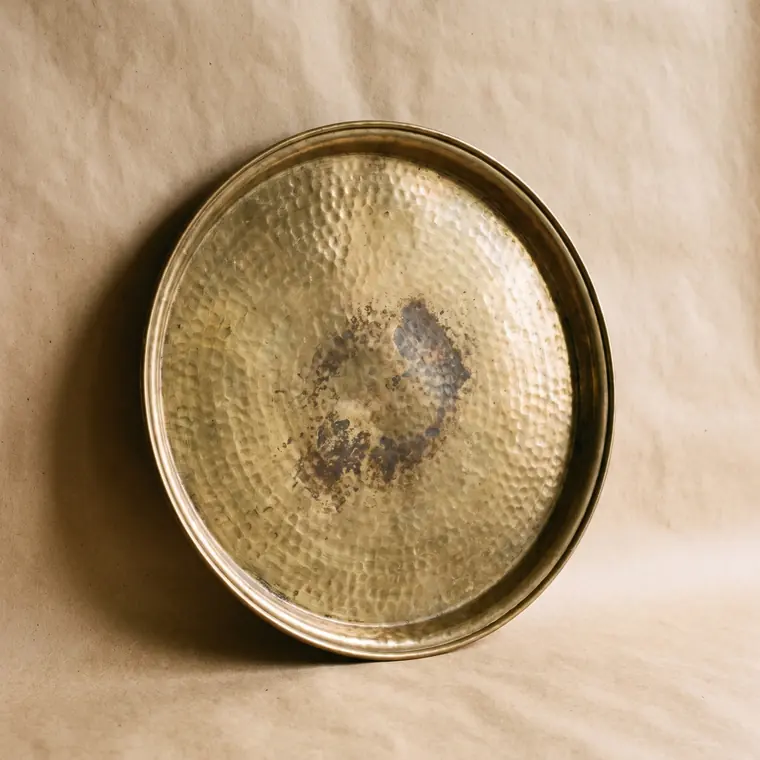 A close view of a small hammered brass dish with warm patina on a warm paper sweep — upper detail.