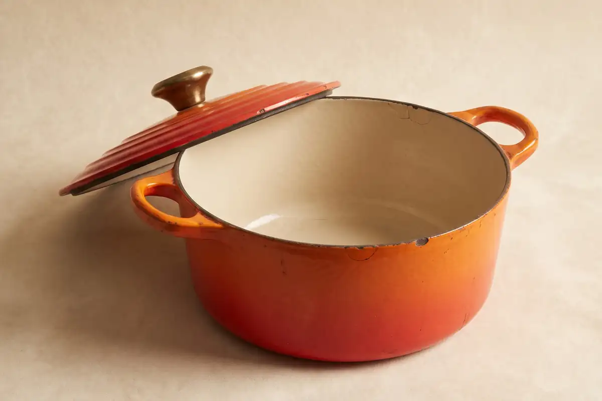 A vintage flame-orange enamelled cast iron casserole with a cream interior and a brass knob on a warm paper sweep — full width on the paper sweep.