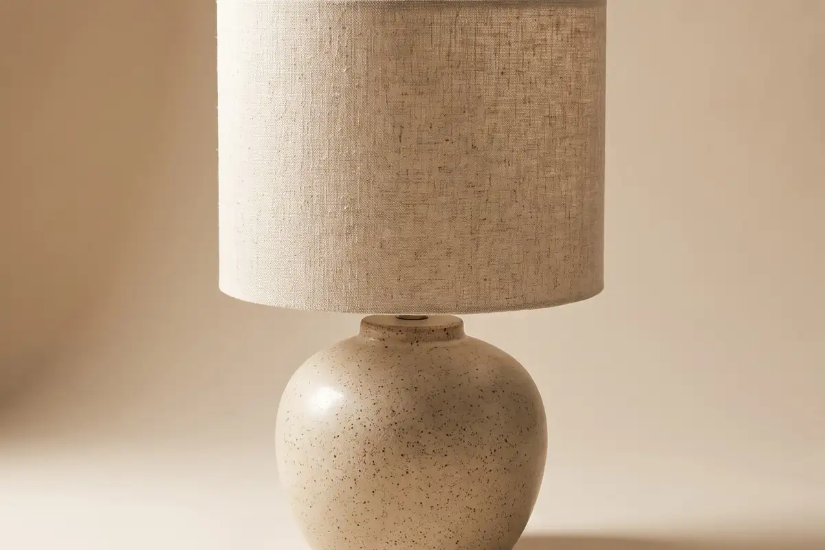 A ceramic table lamp with a fat speckled oatmeal stoneware base and a natural raw linen drum shade on a warm paper sweep — full width on the paper sweep.
