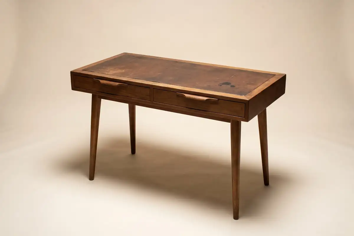 A small mid-century walnut writing desk with two slim drawers and a worn leather top on a warm paper sweep — full width on the paper sweep.