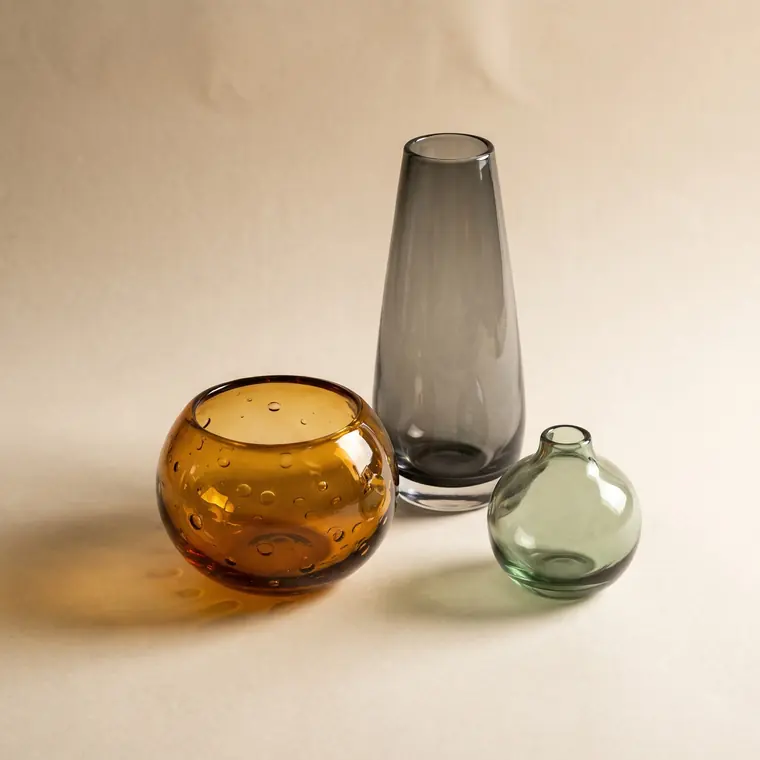 Three hand-blown studio glass vessels in smoke grey, amber and sage, casting warm coloured light on a warm paper sweep — upper detail.
