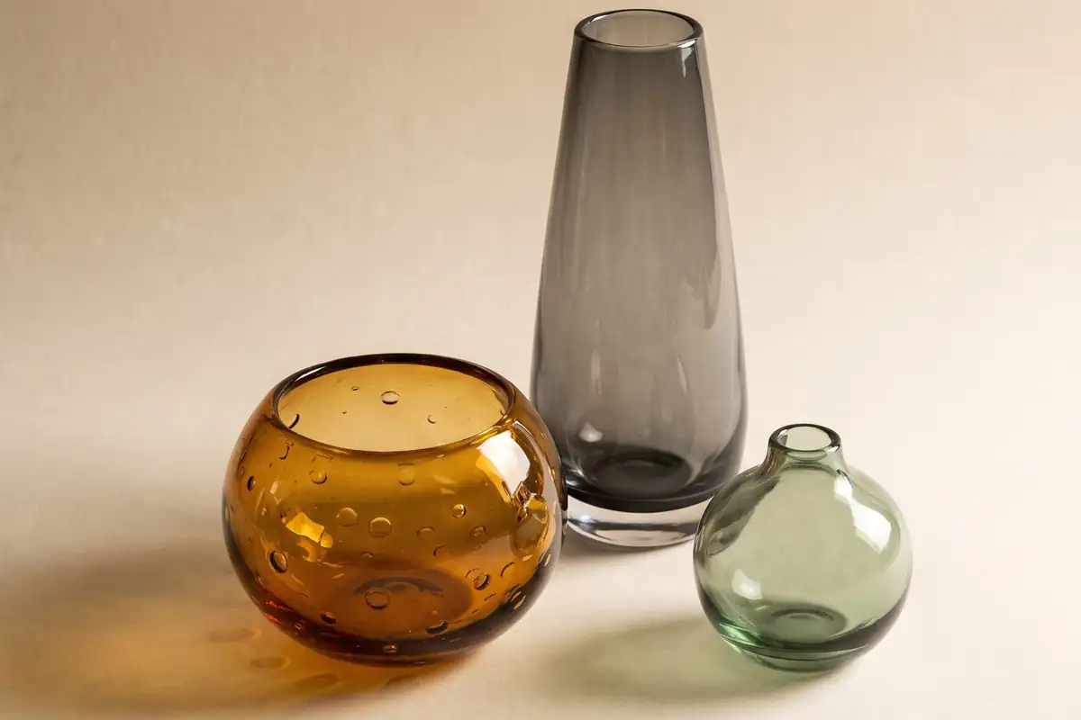 Three hand-blown studio glass vessels in smoke grey, amber and sage, casting warm coloured light on a warm paper sweep — full width on the paper sweep.