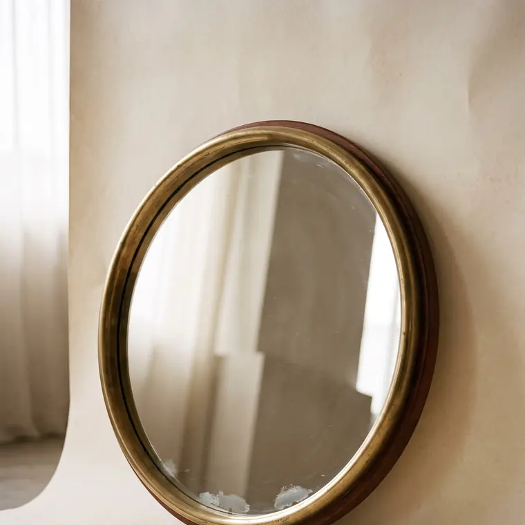 A round mid-century wall mirror with a brass rim and a slim teak surround leaning against a warm paper sweep — upper detail.