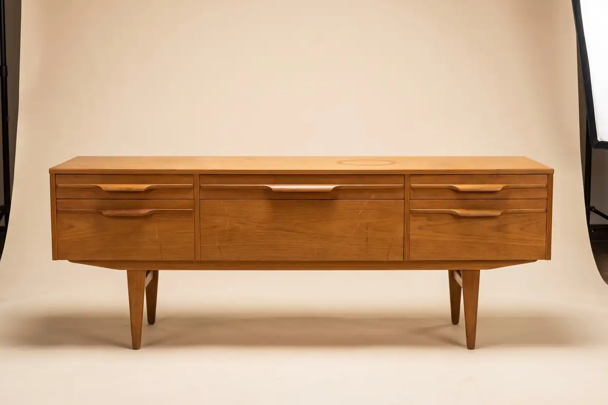 A long low Danish teak sideboard with sculpted drawer pulls and tapered legs against a warm paper backdrop — full width on the paper sweep.