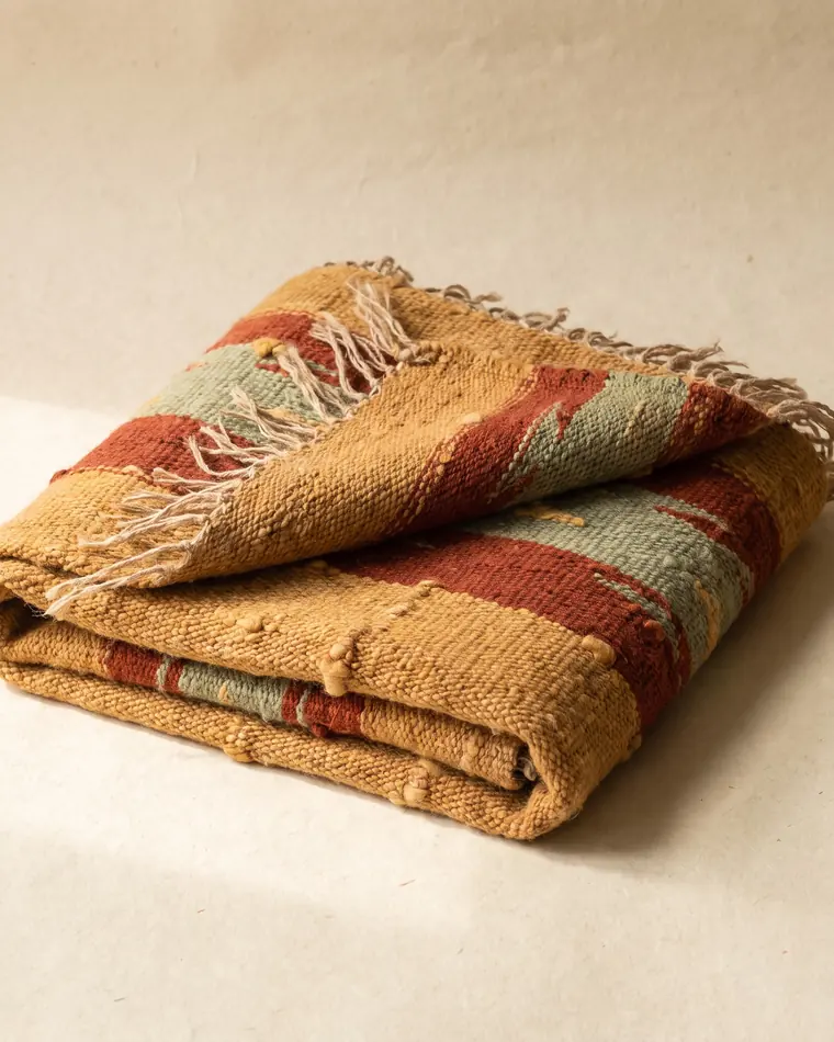 A handwoven wool textile in warm ochre, rust and sage geometric bands, folded with one corner turned to show the reverse.