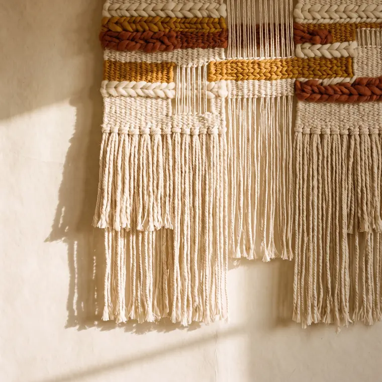 A handwoven wall hanging in undyed wool with ochre and rust bands and long knotted fringe on an oak dowel — lower detail, showing the foot and the shadow.