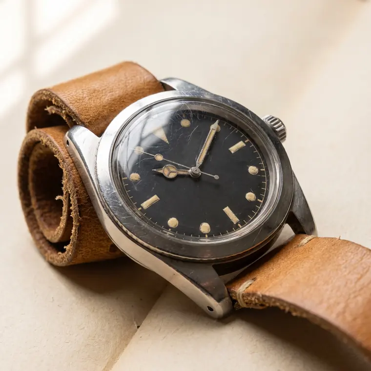 A vintage steel diver’s watch with a faded matte black dial, cream lume plots and a worn tan leather strap on warm paper — upper detail.