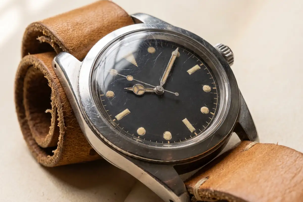 A vintage steel diver’s watch with a faded matte black dial, cream lume plots and a worn tan leather strap on warm paper — full width on the paper sweep.